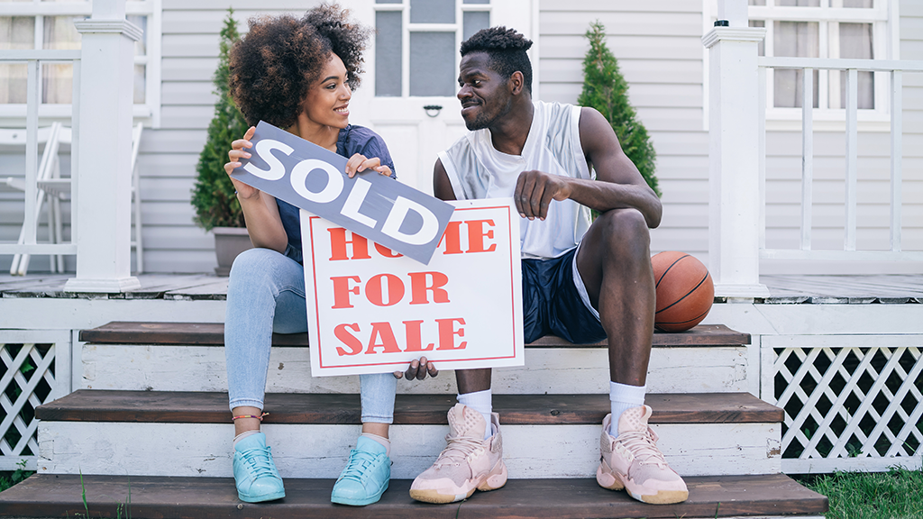 A couple sits on steps to a house with two signs. One sign reads "home for sale" and the other reads "sold."