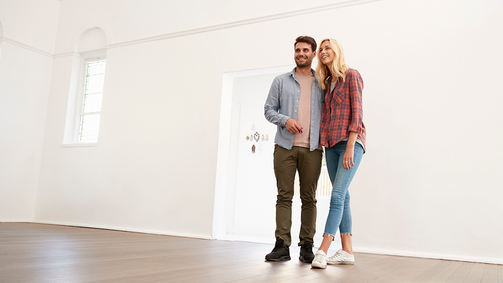 A young couple stands in a bright, empty room with keys in their hands